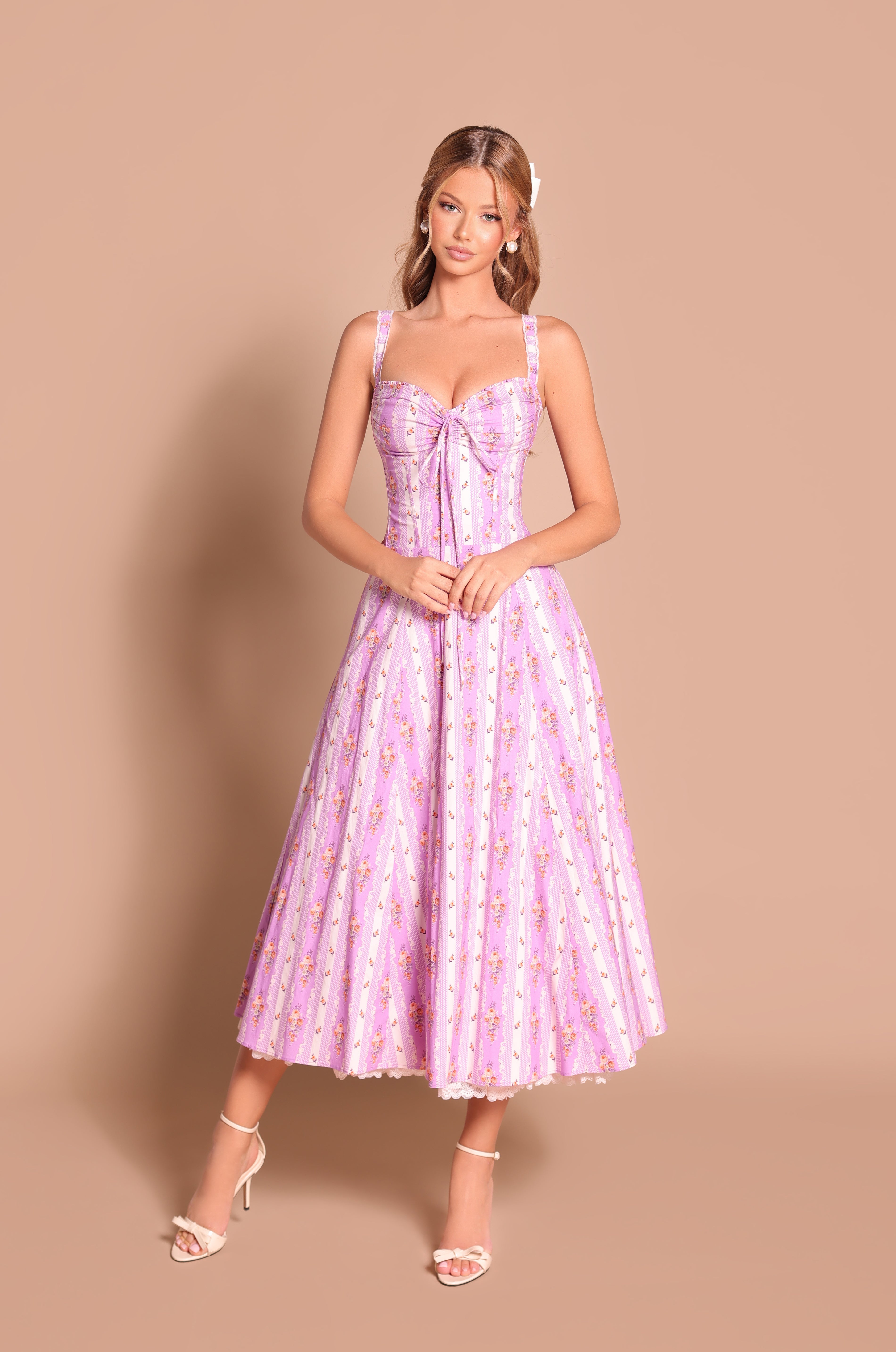 Woman wearing the Celenia Lilac Floral Casual Midi Dress with a pleated skirt and spaghetti straps, paired with white heels, stands against a beige backdrop—a perfect choice for summer weddings.